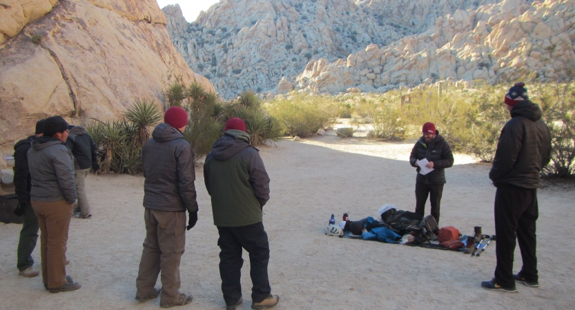 A group of people stand at the base of a large rock formation in a desert environment. They are listening to someone speak. 